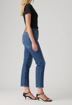 Frau steht seitlich, trägt ein schwarzes eng anliegendes T-Shirt, hoch taillierte blaue Jeans und schwarze Riemchensandalen mit niedrigem Absatz vor schlichtem Hintergrund.