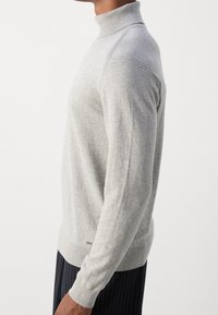 Lysegrå rullekrave-trøje med ribbet krave og kant, med ensartet tekstur og tætsiddende pasform, båret med mørke bukser med tynde striber.