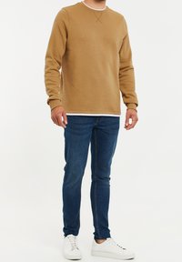 Sudadera marrón con cuello redondo, tejido texturizado y un detalle blanco en el dobladillo, combinada con jeans ajustados azules y zapatillas blancas.