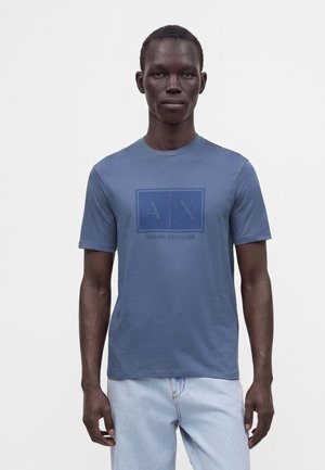 Młody mężczyzna ubrany w niebieską koszulkę Armani Exchange z logo na piersi oraz jasnoniebieskie dżinsy na jednolitym białym tle.