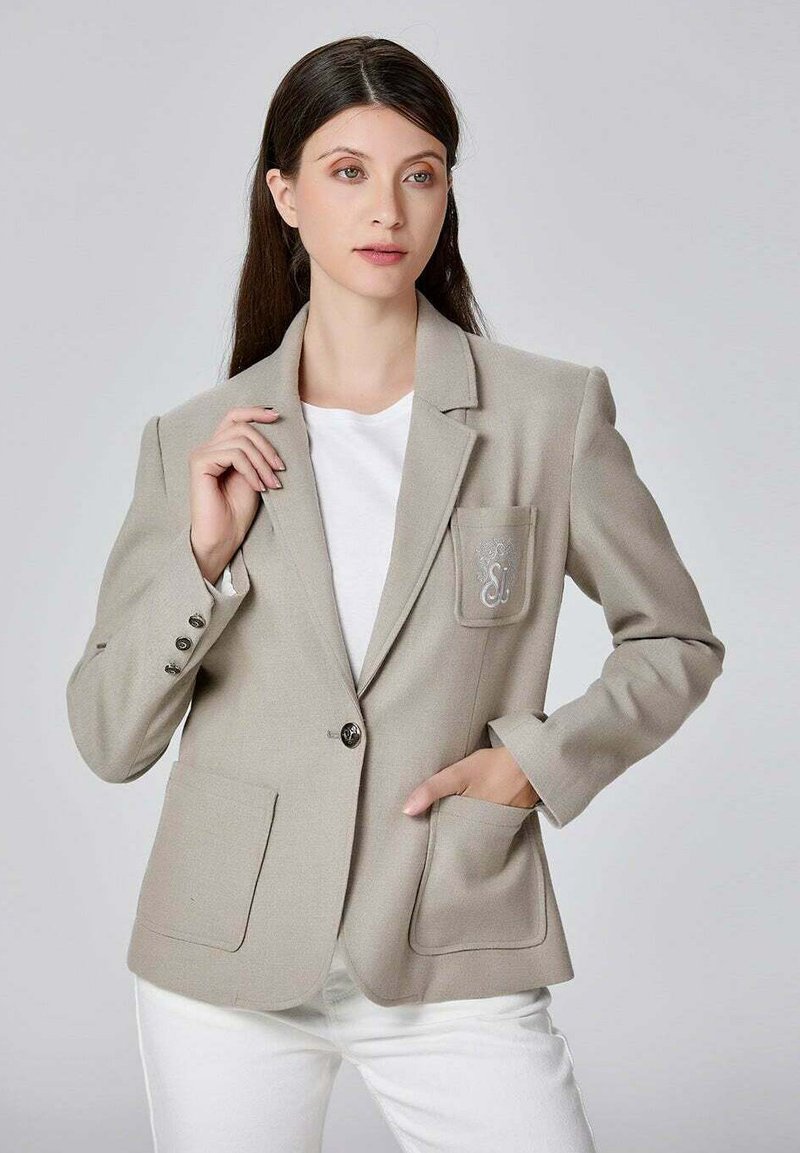 Blazer beige en tissu texturé, doté d'une fermeture à un bouton, de deux poches avant, et d'une poche avec un détail brodé.