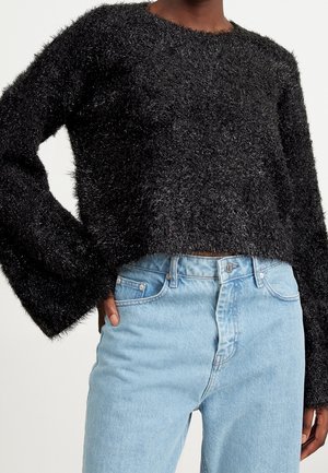 Personne portant un pull noir duveteux à manches longues et un jean taille haute bleu clair, une main dans la poche.