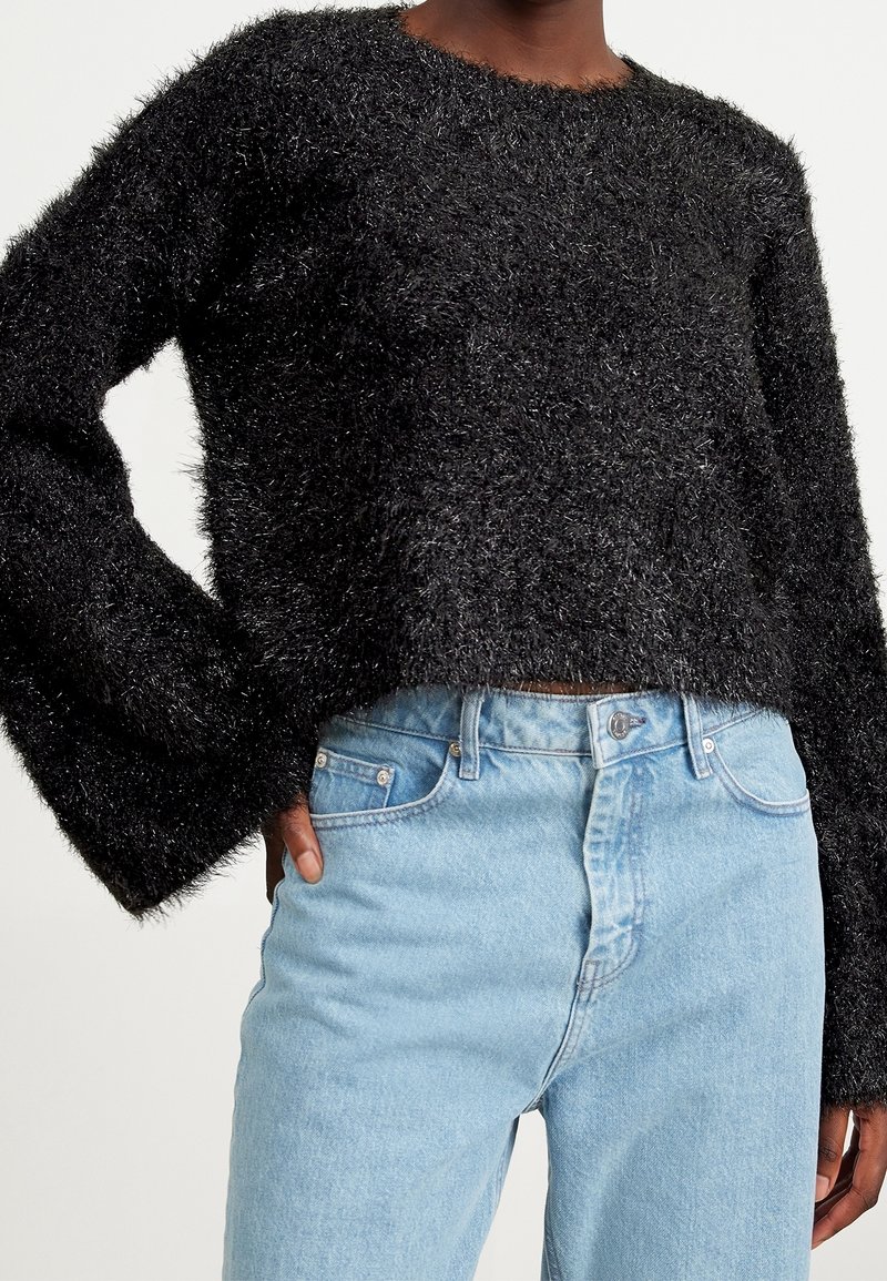 Personne portant un pull noir duveteux à manches longues et un jean taille haute bleu clair, une main dans la poche.