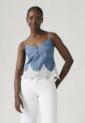 Mujer con top de mezclilla azul sin mangas con borde de encaje blanco y pantalones blancos, de pie contra un fondo gris claro.