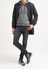 Cappotto nero leggero, felpa grigia con coulisse, jeans scuri e sneakers nero-grigie. Texture lisce e design casual.