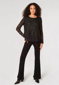 Zwarte kanten top met lange mouwen en pailletten, gelaagd over een zwarte doorschijnende blouse; gecombineerd met zwarte geribde flared broeken en puntige enkellaarzen.