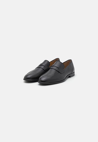 Black leather loafers with a smooth texture, rounded toe, and a decorative strap across the upper. Flat sole, minimal design.