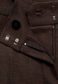 Hnědé látkové kalhoty s černým knoflíkem, kovovými detaily a kapsou na zip. Texturovaný materiál s viditelnými švy.