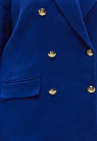 Plavi dvostruko kopčani sako sa ribljim kosturom, teksturiranim tkanjem, zlatnim ukrasnim gumbima i jednim džepom na lijevoj strani.