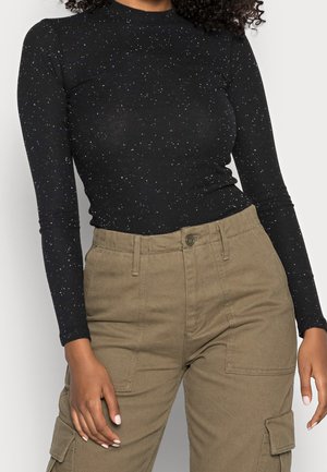 Vrouw draagt een aansluitende zwarte gestippelde top met lange mouwen en een high-waisted kaki cargobroek met zakken en knoopsluiting.