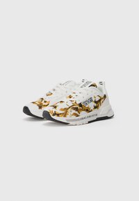 White sneakers with gold baroque print, black soles, and "Versace Jeans Couture" branding on sides and tongue.