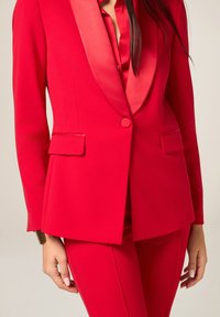 Czerwona marynarka z atłasowymi klapami, zapinana na jeden guzik, z dwoma przednimi kieszeniami, zestawiona z pasującymi czerwonymi spodniami, gładka tekstura tkaniny.