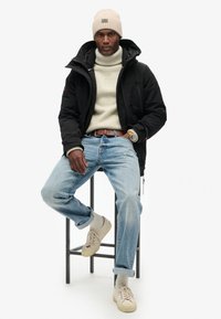 Chaqueta de plumas negra, suéter de cuello alto color crema, jeans azul claro, zapatillas beige y un gorro de lana beige; sentado en un taburete negro.