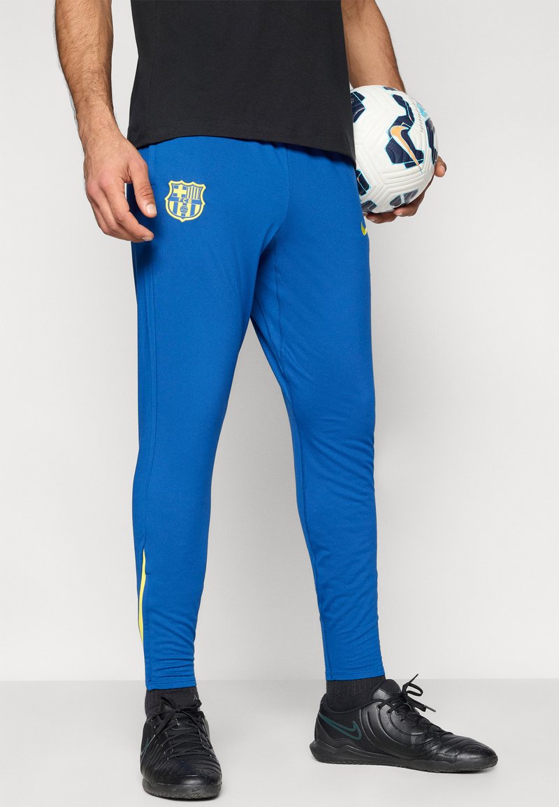 Blaue Sporthose mit tailliertem Schnitt, versehen mit einem gelben Logo, Seitenstreifen und einem glatten, leichten Stoff. Kombiniert mit schwarzen Fußballschuhen.