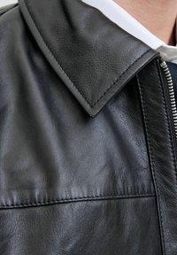 Schwarze Lederjacke mit spitzem Kragen, strukturiertem Oberflächenmaterial, Reißverschluss und sichtbaren Nähdetails. Nahaufnahme des Stoffes des Kleidungsstücks.