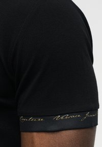 Κοντινό πλάνο μαύρου υφασμάτινου κοντομάνικου με ριμπ μανσέτα που φέρει χρυσά καλλιγραφικά γράμματα στο χέρι ενός ατόμου.