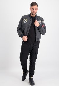 Giubbotto bomber nero con toppe NASA, chiusura con zip, polsini e orlo a costina. Indossato sopra una maglietta nera e abbinato a pantaloni cargo neri e scarpe da ginnastica.