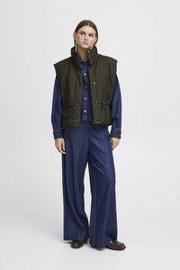 Roheline tikandiga vest kõrge kaelusega, reguleeritavad paelad vööl ja nööbid sinise teksajaki ning laia säärega siniste pükste peal.