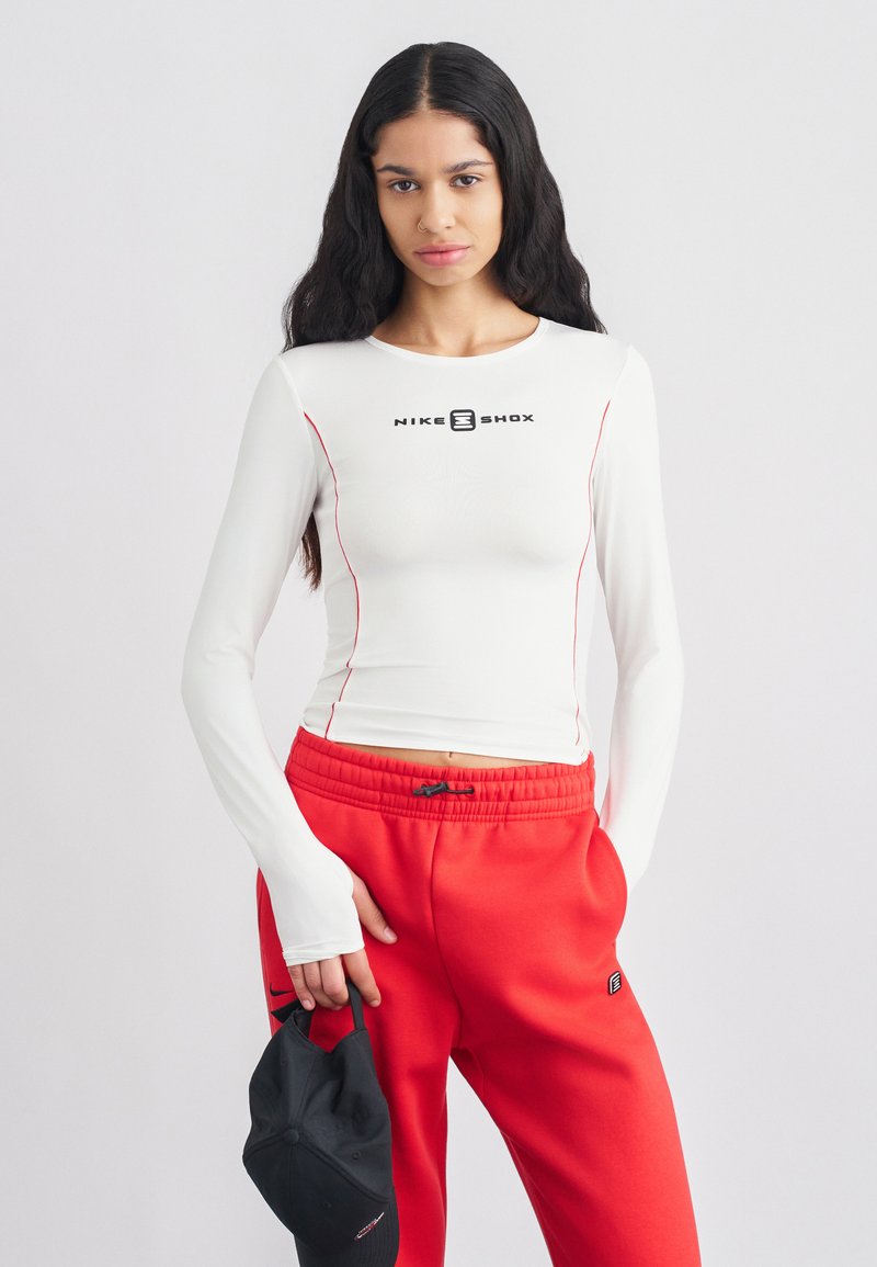 Bijela majica s dugim rukavima s crvenim detaljima, s crnim logotipom. Uparena s crvenim hlačama i crnom kapom, prikazujući sportski dizajn.