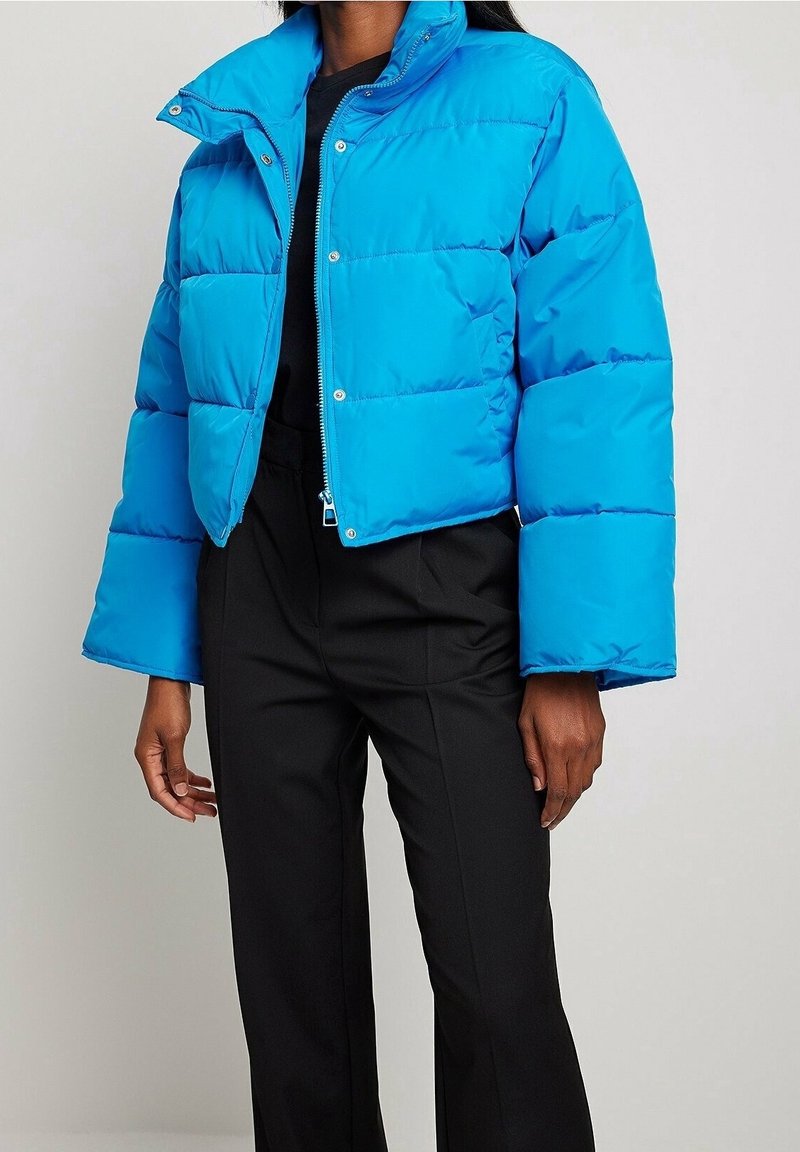 Chaqueta de plumas azul con un diseño corto, que presenta un cuello alto, cierre de cremallera y patrones de acolchado horizontales. Combinada con pantalones negros.