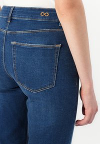 Dark blue denim jeans, featuring a fitted shape, golden stitching, and a subtle infinity symbol detail on the back waistband.