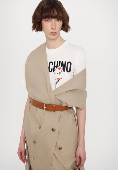 Beige, Wickelkleid mit großen Knöpfen und einem braunen Ledergürtel. Darüber trägt es ein weißes T-Shirt mit grafiklichem Vogel-Design.