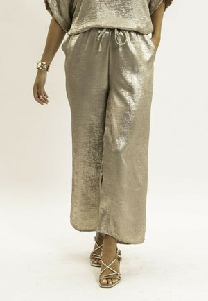 Femme portant un pantalon large brillant doré et des sandales à talons hauts assorties à fines lanières, avec un bracelet doré au poignet gauche.