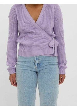 Lila gebreide wikkeltrui met lange mouwen, V-hals en een zijstrikdetail, gecombineerd met lichtblauwe high-waisted jeans.