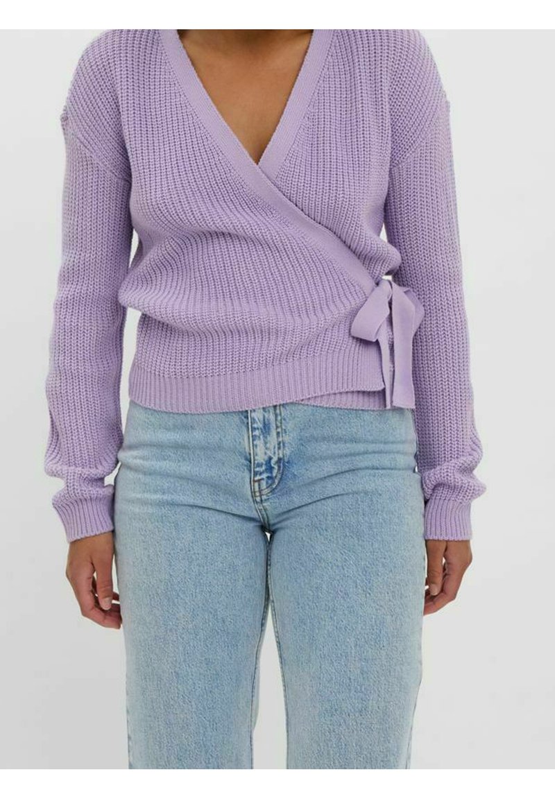 Fioletowy sweter z dzianiny w formie narzutki, z długimi rękawami, dekoltem w kształcie litery V oraz bocznym wiązaniem, zestawiony z jasnoniebieskimi spodniami o wysokim stanie.