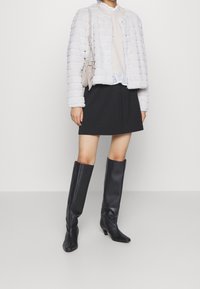 Damenoutfit mit einer strukturierten weißen cropped Jacke, beigen Pullover, schwarzem Rock und knielangen schwarzen Lederstiefeln.