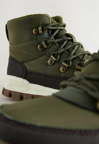 Grüner wasserdichter Stiefel mit textilem Obermaterial, metallenen Ösen und einer dicken Gummisohle mit strukturiertem Finish. Akzente in Dunkelbraun.