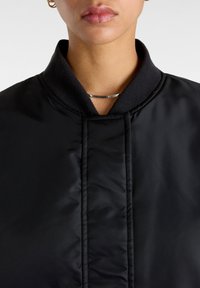Veste matelassée noire avec col montant et accent métal argenté au niveau de l'encolure. Texture lisse avec des détails de couture subtils.