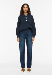 Maglione a coste color navy con collo a camicia e chiusura con cinque bottoni, abbinato a jeans a gamba dritta blu scuro a vita alta e scarpe con tacco a blocco color cuoio.