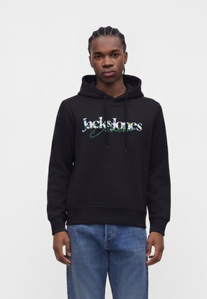 Jeune homme aux cheveux tressés portant un sweat à capuche noir Jack & Jones et un jean bleu, debout devant un fond clair uni.