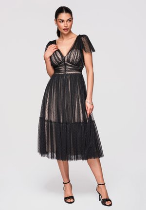 Kvinde iført en tynd sort polkaprikket tulle-midi-kjole med kappelærmer og sorte højhælede sandaler, poserer mod en ensfarvet baggrund.