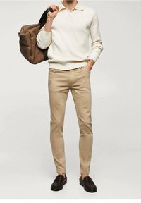 Pantalones estrechos de color beige claro combinados con un suéter blanco texturizado y zapatos de piel marrón tipo mocasín. Se sostiene un bolso marrón con un bolsillo frontal.