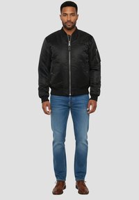 Zwarte bomberjack met ritssluiting aan de voorkant, geribbelde manchetten en tailleband. Draagt lichtblauwe spijkerbroek en bruine schoenen, staand tegen een grijze achtergrond.