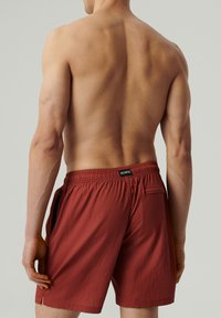 Czerwone szorty kąpielowe z teksturowanego materiału, z elastycznym paskiem w talii, bocznymi kieszeniami i małą kieszenią z tyłu. Widoczna etykieta marki z tyłu.