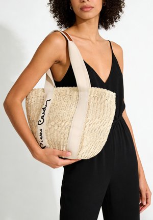 Donna che indossa una tuta nera senza maniche che tiene in mano una borsa tote beige intrecciata con ampie cinghie crema e dettagli in scrittura nera.