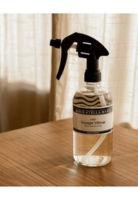 Bottiglia di spray per ambienti etichettata "Voyage Vétiver" con un nebulizzatore nero su una superficie di legno, con tende trasparenti sullo sfondo.