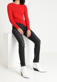 Röd ribbad långärmad tröja kombinerad med åtsittande svarta jeans och vita kilklackade stövlar på en vit pelare mot en neutral bakgrund.
