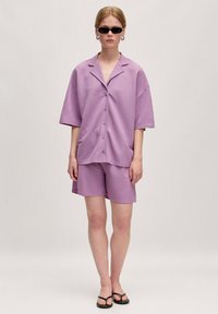 Lichtpaarse shorts en een oversized button-up shirt van zacht materiaal. De outfit heeft een relaxte pasvorm en korte mouwen. Zwarte zonnebril gedragen.
