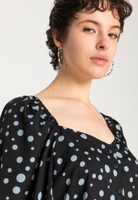 Blusa preta com poás azul claro, decote quadrado, mangas bufantes e fecho frontal com botões. Tecido suave com ligeiro brilho.