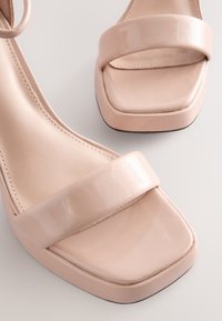 Blush laklæder høje hæle med firkantet tå, brede remme foran og et slankt design. Glat tekstur og minimale hardware-detaljer.