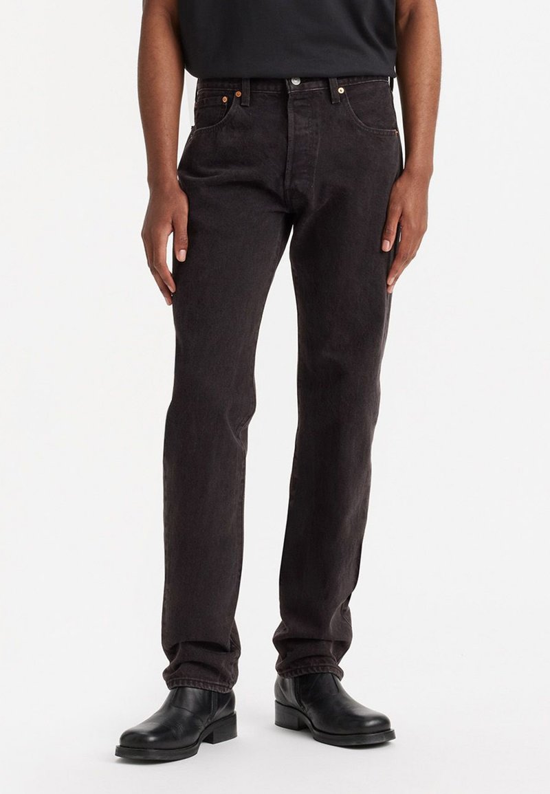 Persona que lleva puestos unos jeans negros de pierna recta con botón y cremallera, combinados con unas botas de cuero negras hasta el tobillo y una camiseta negra.