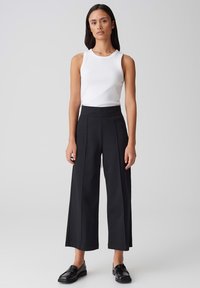 Pantalons larges noirs à taille haute, avec deux plis à l'avant. Associés à un débardeur blanc ajusté et des mocassins noirs. Texture lisse.