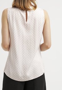 Hihaton beige paita, jossa on hillitty geometrinen kuvio, yllään henkilö, joka on kuvattu takaa, yhdistettynä mustiin housuihin.