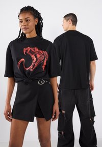 Black t-shirt with red snake graphic, paired with a black wrap skirt featuring an adjustable belt. Model shows loose fit and contemporary design.