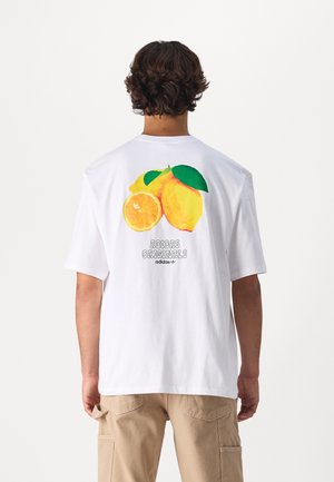 Homme aux cheveux bouclés porte un t-shirt blanc Adidas avec un grand motif citron et orange et le texte « Adidas Originals » au dos.