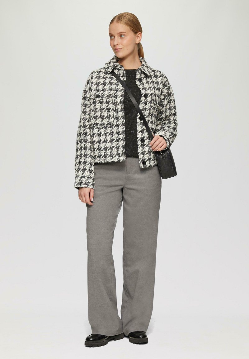 Schwarze und weiße Hahnentrittmuster-Jacke, cropped Länge, Knopfleiste vorne. Schwarz pullover darunter. Graue weite Hose, schwarze Schuhe und eine kleine schwarze Tasche.